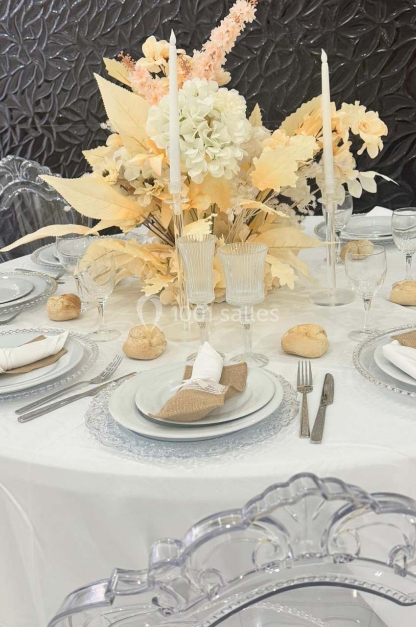 Table élégamment dressée avec vaisselle transparente, bougies blanches et centre de table floral beige et blanc.