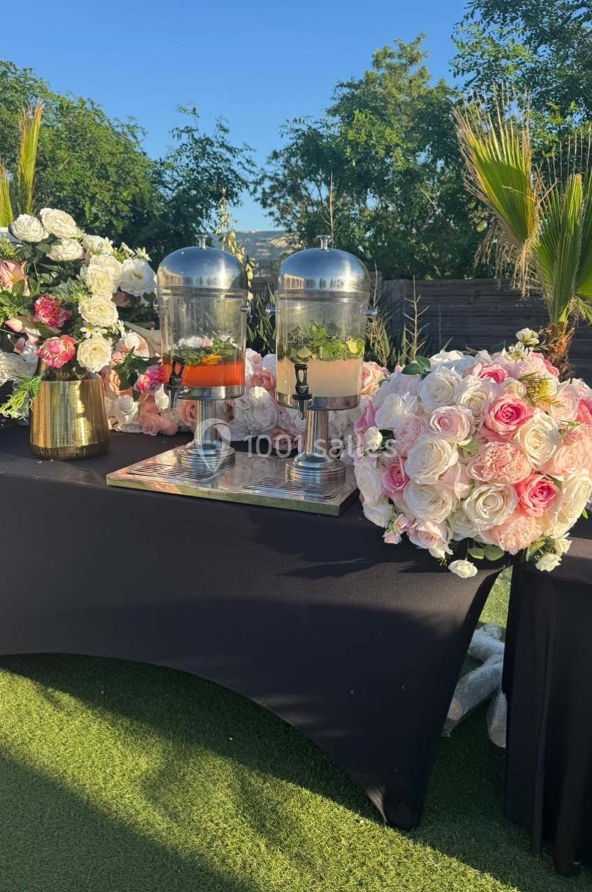 Distributeurs de boissons sur une table noire décorée de fleurs roses et blanches, en extérieur par temps ensoleillé.