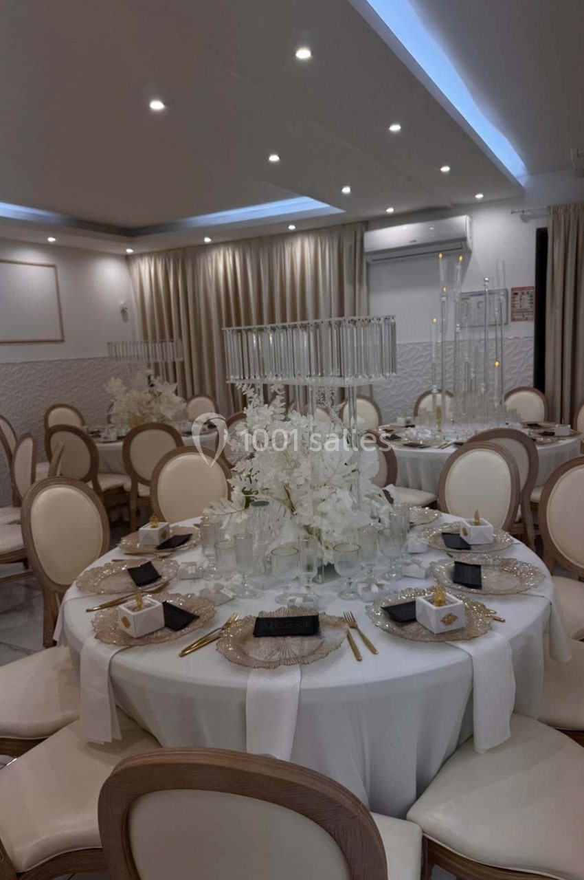 Salle de réception élégante avec tables rondes décorées de nappes blanches, chandeliers et arrangements floraux blancs.