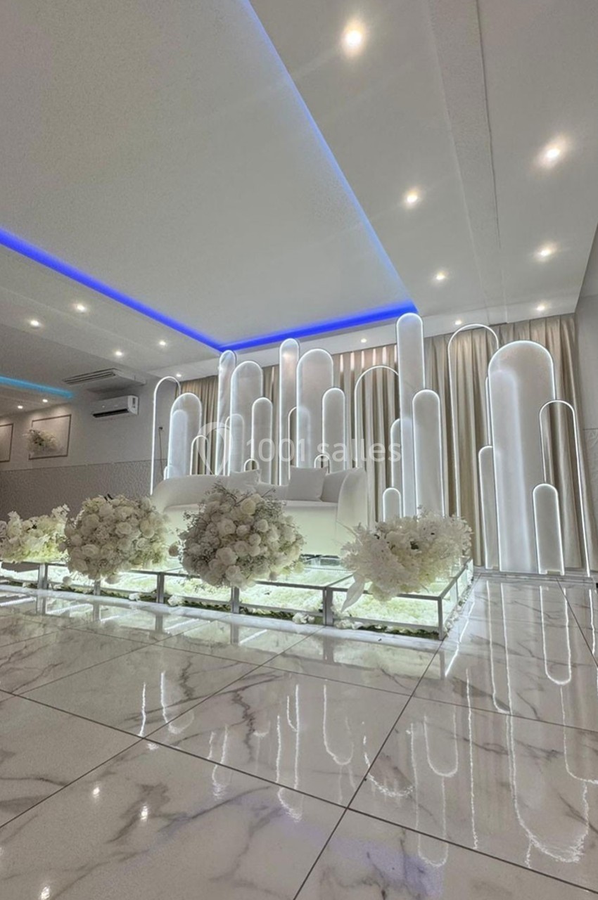 Décor intérieur élégant avec podium blanc, colonnes lumineuses et arrangements floraux sur sol en marbre.