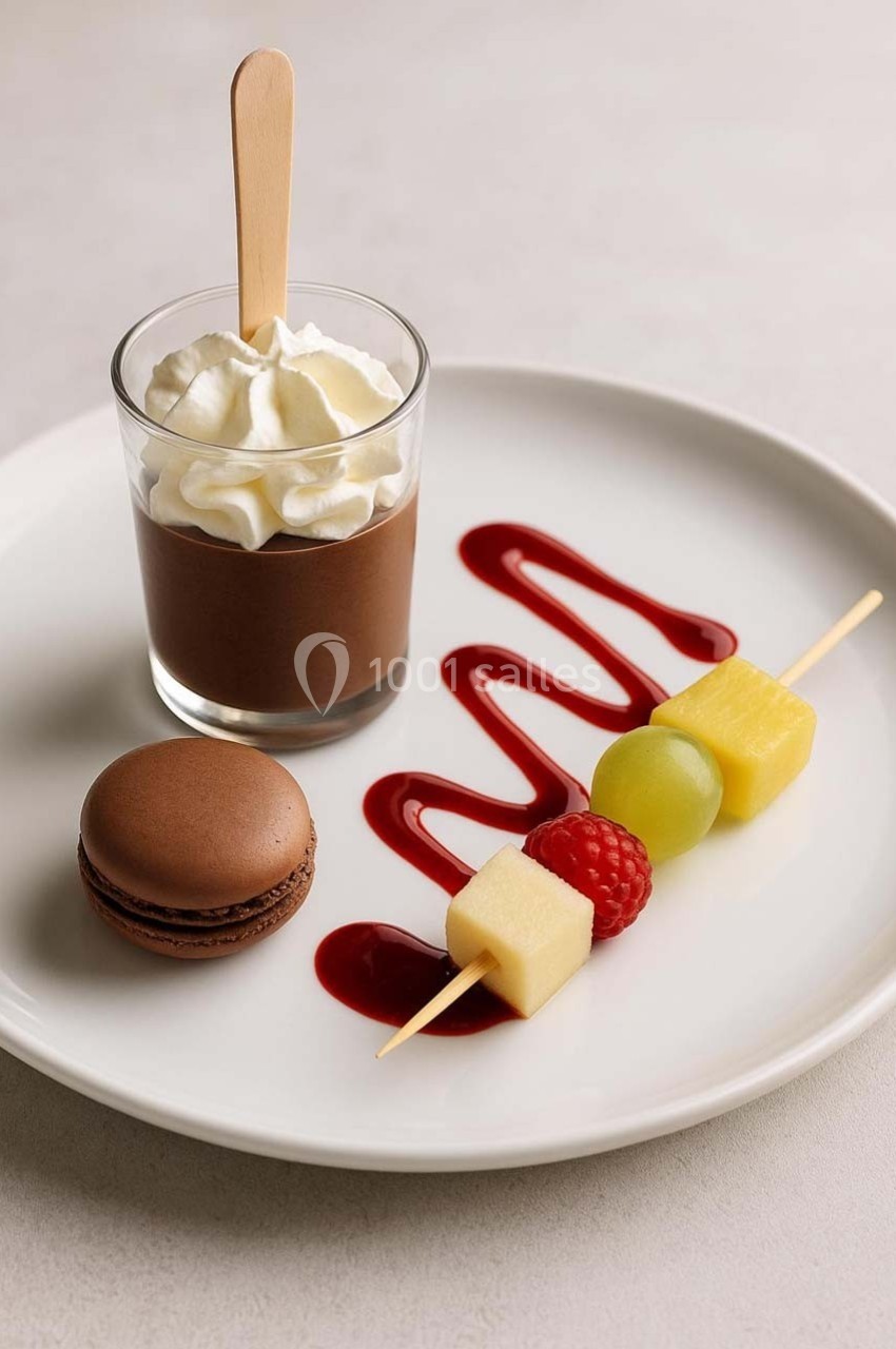 Dessert composé d'une mousse au chocolat, d'un macaron, de fruits sur brochette et d'un coulis rouge sur une assiette…