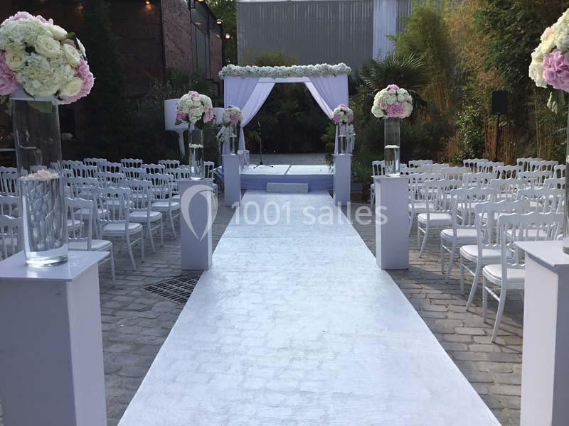 Allée centrale décorée de fleurs menant à une arche blanche pour une cérémonie en extérieur, entourée de chaises alignées.