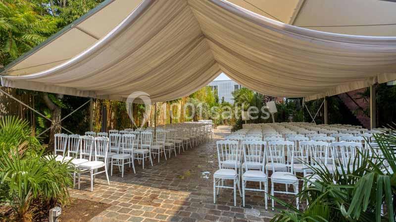 Chaises blanches alignées sous une tente élégante dans un espace extérieur pavé entouré de végétation.
