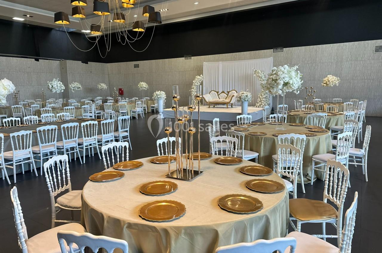 Salle de réception élégante avec tables rondes dressées de nappes dorées, chaises blanches et décor floral blanc.