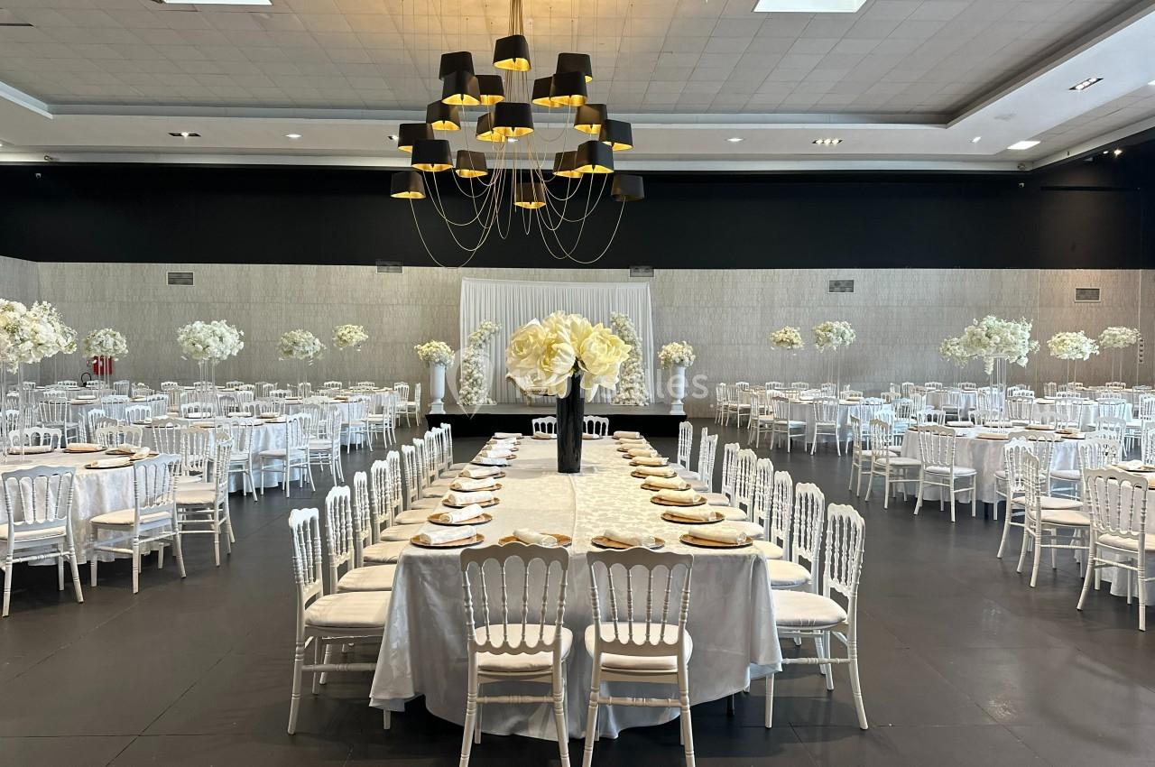 Salle de réception élégante avec tables dressées, chaises blanches et décoration florale blanche sous un grand lustre noir.