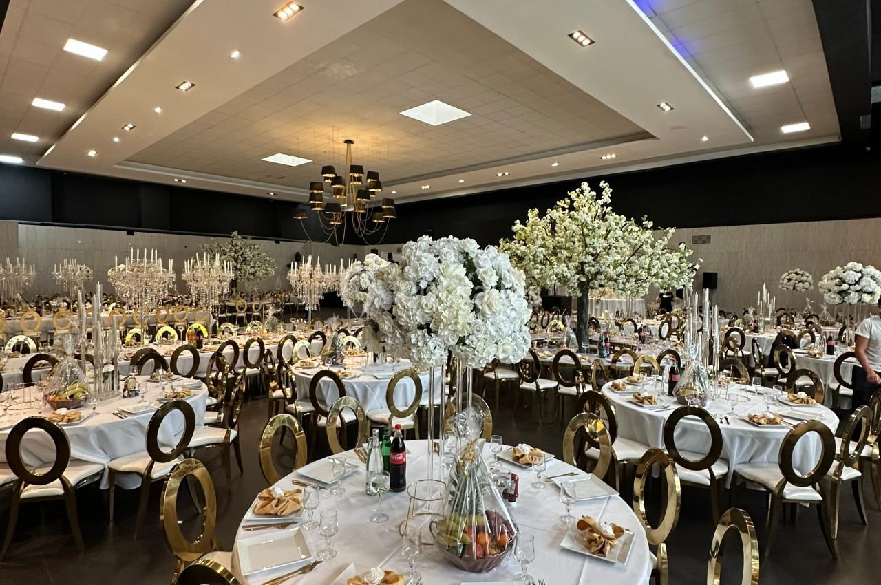 Salle de réception élégante avec tables rondes décorées de nappes blanches, chaises dorées et centres de table fleuris.