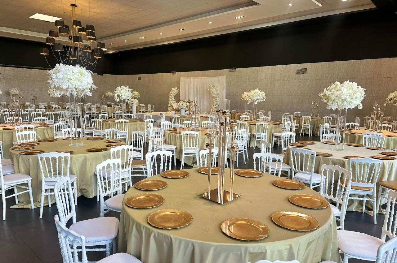 Salle de réception décorée avec des tables rondes, nappes dorées, assiettes dorées et centres de table fleuris blancs.