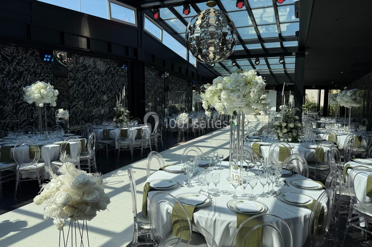 Salle de réception élégante avec tables rondes dressées, décorations florales blanches et plafond vitré.
