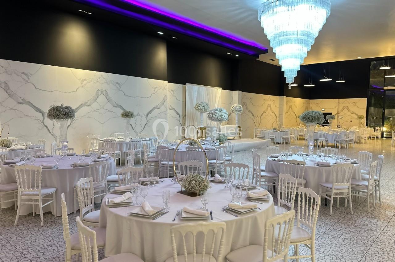 Salle de réception élégante avec tables rondes dressées, chaises blanches et grand lustre lumineux.