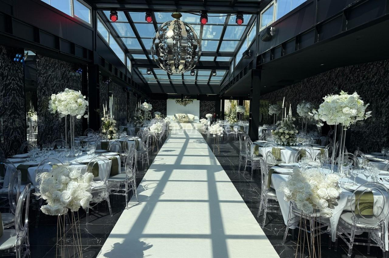 Salle de réception élégante avec tables décorées de fleurs blanches et allée centrale sous un plafond en verre.