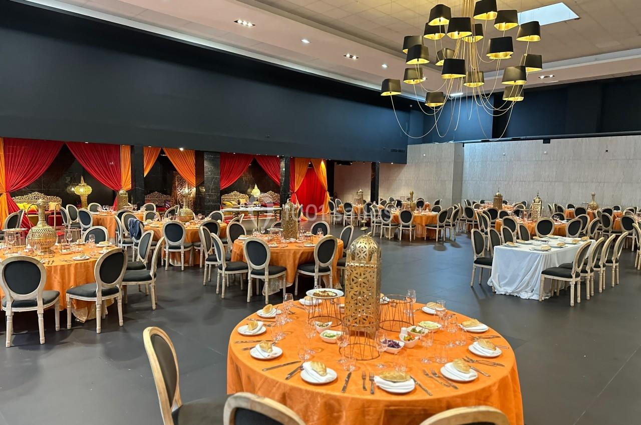 Salle de réception décorée avec des tables rondes nappées d'orange, des chaises noires et un grand lustre moderne.