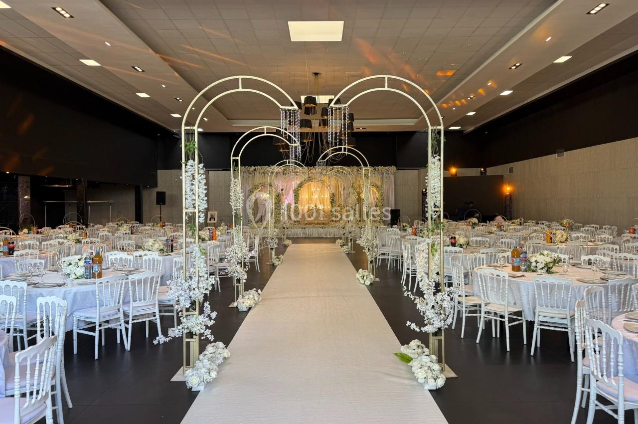 Salle de réception décorée avec des tables rondes, des chaises blanches et une allée centrale ornée d'arcs fleuris.