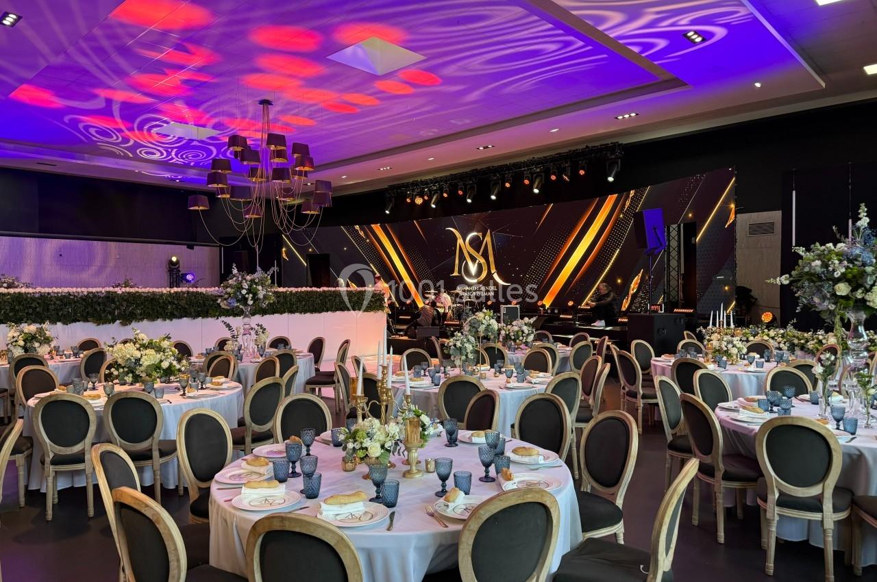 Salle de réception élégante avec tables décorées, éclairage coloré et scène équipée pour un événement festif.