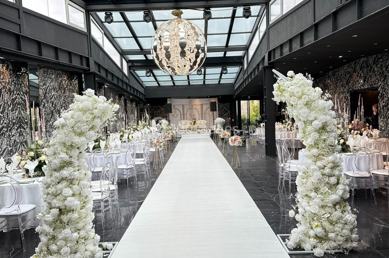 Allée centrale d'une salle de réception décorée de fleurs blanches, avec un plafond en verre et un lustre suspendu.