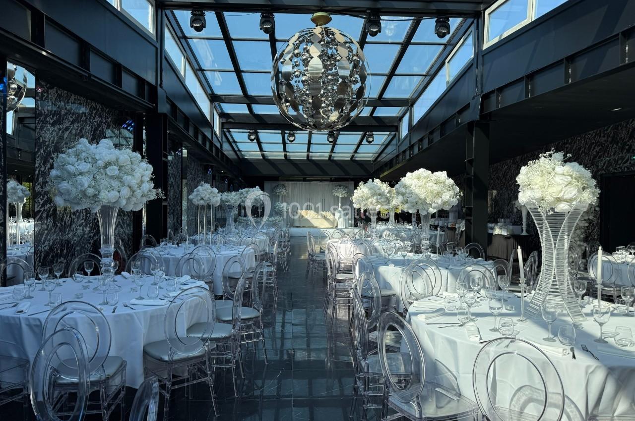 Salle de réception élégante avec tables rondes dressées, chaises transparentes et décorations florales blanches.