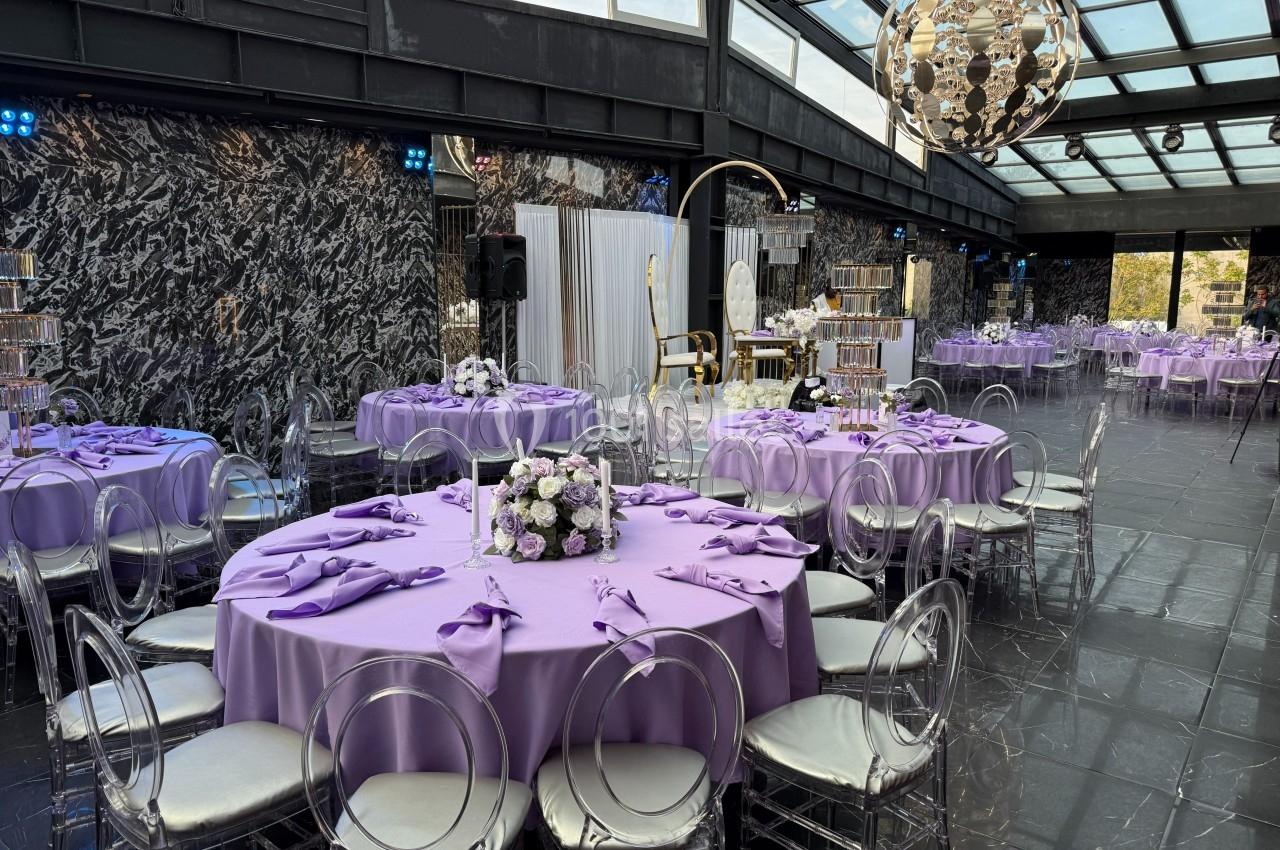 Salle de réception élégante avec tables rondes dressées de nappes violettes, chaises transparentes et décor floral.