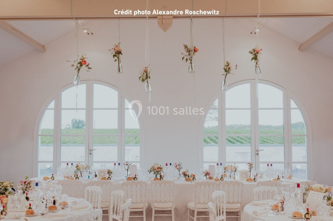 Salle de réception lumineuse avec tables décorées et fleurs suspendues, grandes fenêtres donnant sur un paysage champêtre.