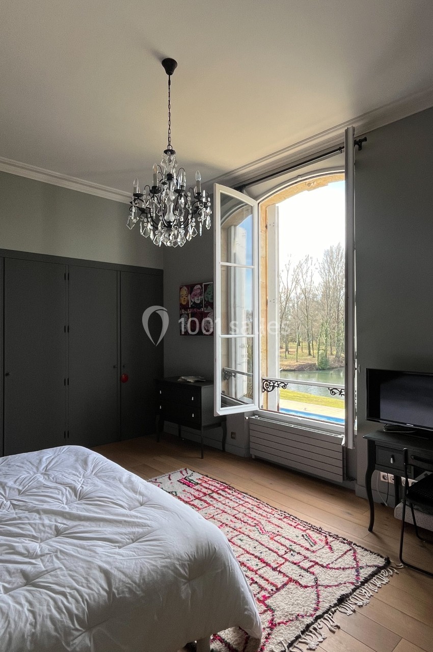 Chambre lumineuse avec lit, armoire grise, lustre, tapis coloré et fenêtre ouverte donnant sur un parc arboré.