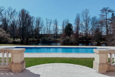 Miniature Piscine du Château Vue sur une piscine extérieure entourée de pelouse, avec un lac et des arbres en arrière-plan sous un ciel dégagé.