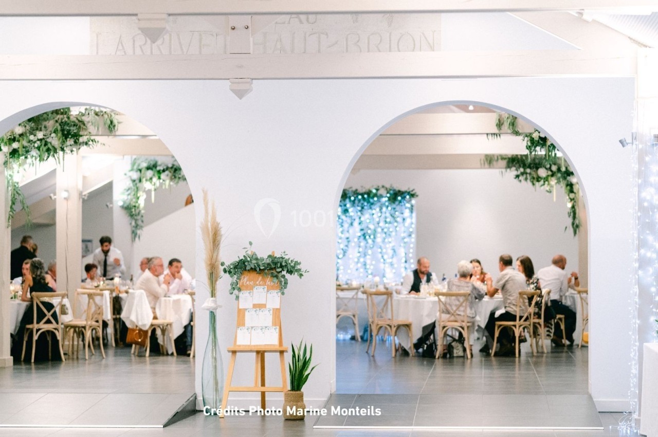 Salle de réception lumineuse avec des tables décorées, des invités attablés et des arches ornées de végétation.