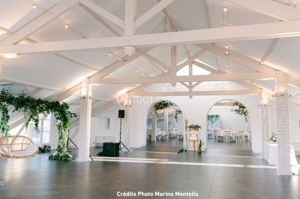 Salle lumineuse avec poutres apparentes blanches, décorée de guirlandes lumineuses et de plantes vertes suspendues.
