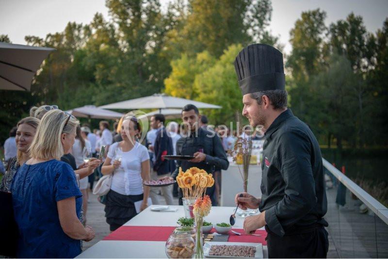 Un chef en tenue noire sert des invités lors d'un événement en plein air près d'un espace arboré.