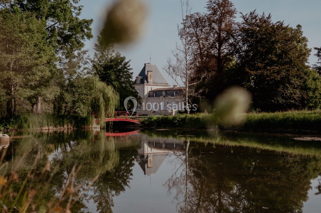 Vue d'un château entouré de végétation, reflété dans un étang avec un pont rouge au premier plan.