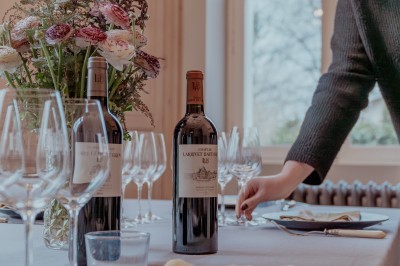 Bouteilles de vin rouge sur une table dressée avec des verres, des fleurs et une personne ajustant un couvert.