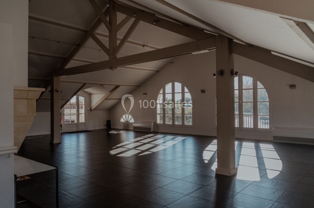 Salle lumineuse sous combles avec poutres apparentes, grandes fenêtres en arche et sol en carrelage sombre.