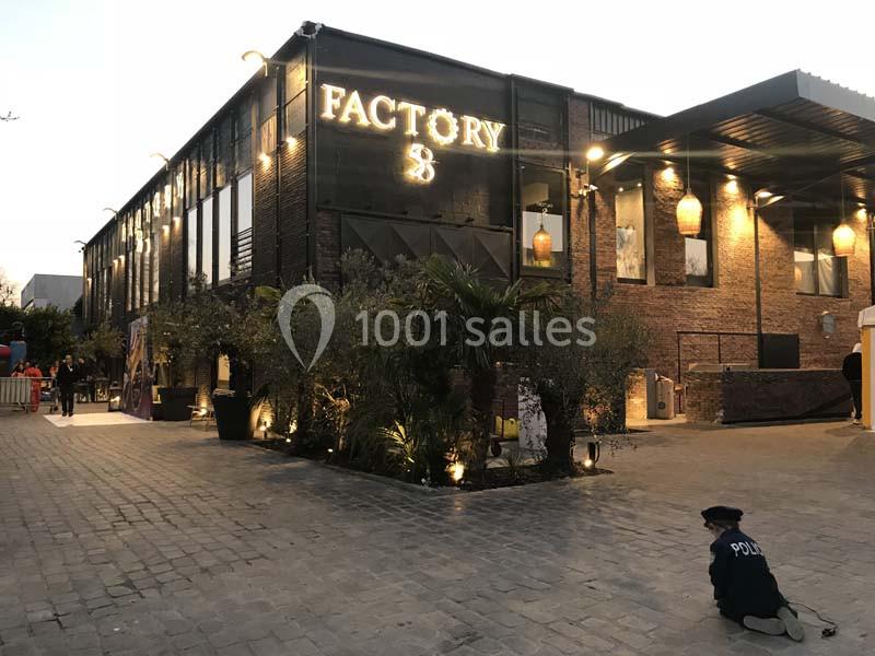 Bâtiment en briques avec enseigne lumineuse ’Factory 8’, entouré de plantes et éclairé en soirée.