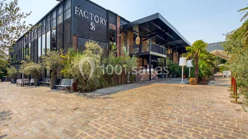 Façade d'un bâtiment industriel moderne entouré de végétation, avec une cour pavée au premier plan.