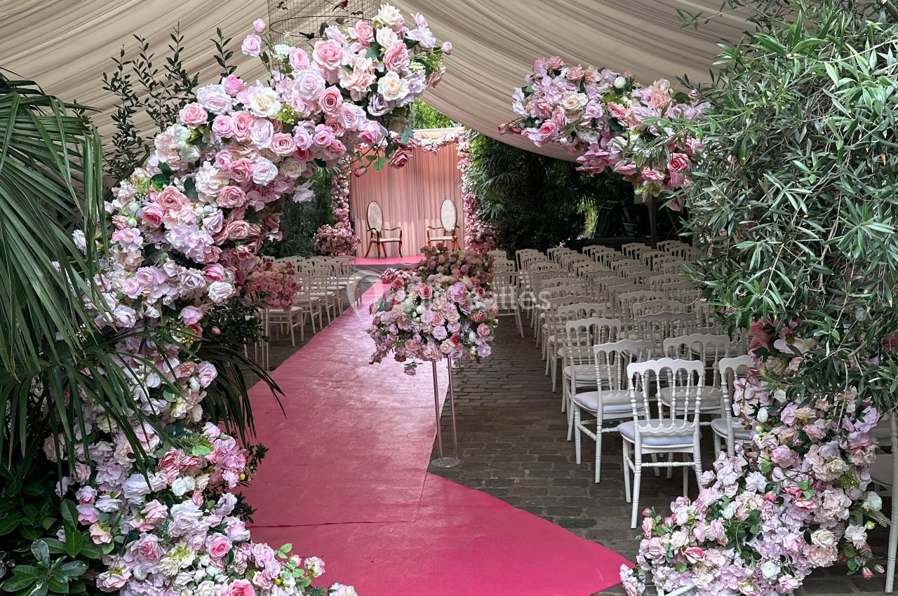 Allée centrale rose bordée d'arcs fleuris sous une tente blanche, avec des chaises alignées pour une cérémonie.