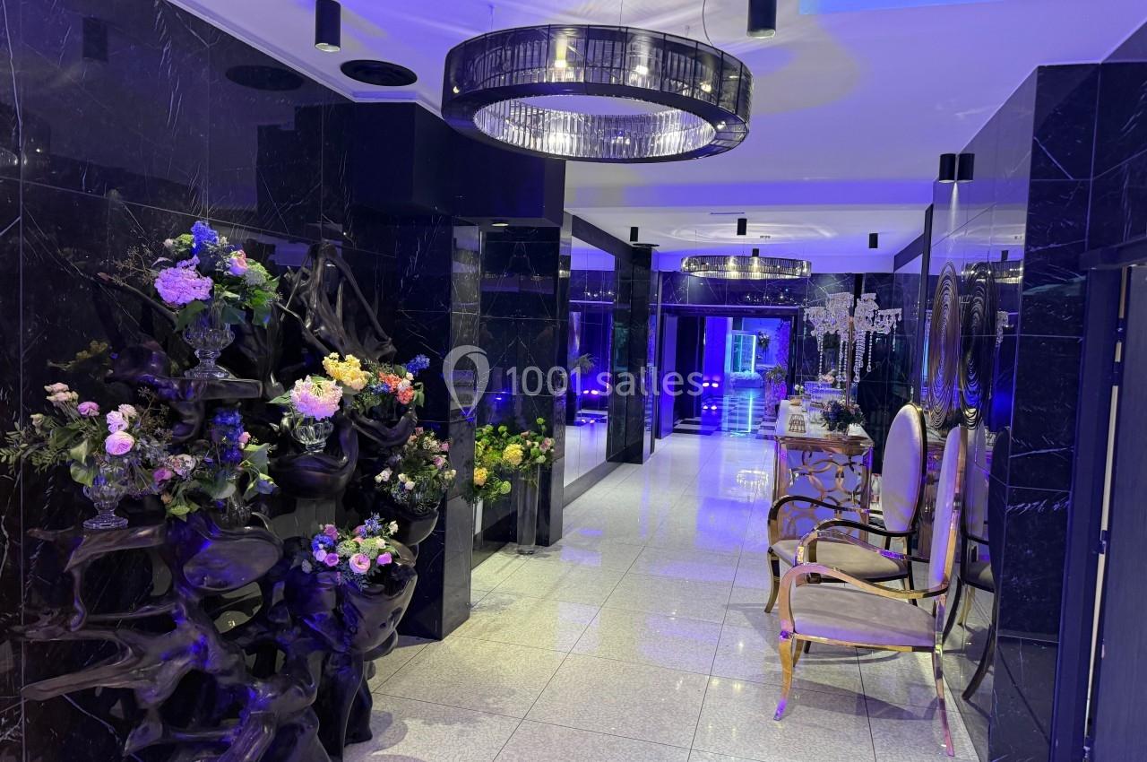 Couloir élégant avec murs noirs brillants, lustres modernes, décorations florales et chaises dorées.