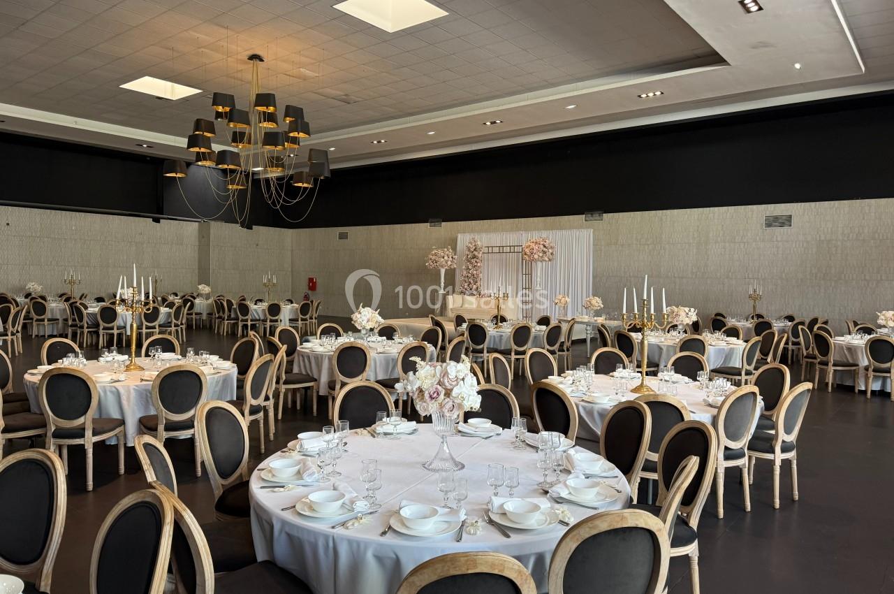 Salle de réception élégante avec tables rondes dressées, chaises noires et décorations florales blanches.