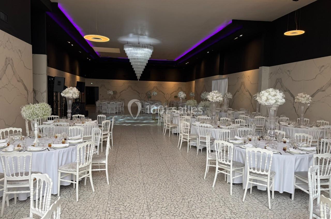 Salle de réception élégante avec tables rondes décorées de fleurs blanches et éclairage violet au plafond.