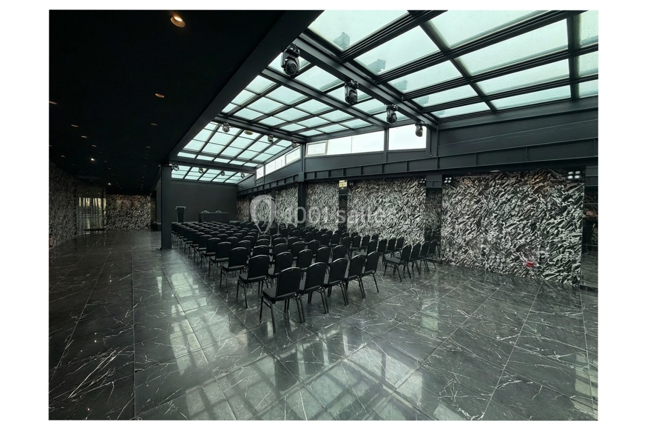 Location salle Bobigny (Seine-Saint-Denis) - Factory 58 #5 Salle de conférence moderne avec des rangées de chaises noires, sol en marbre et toit vitré laissant entrer la lumière…