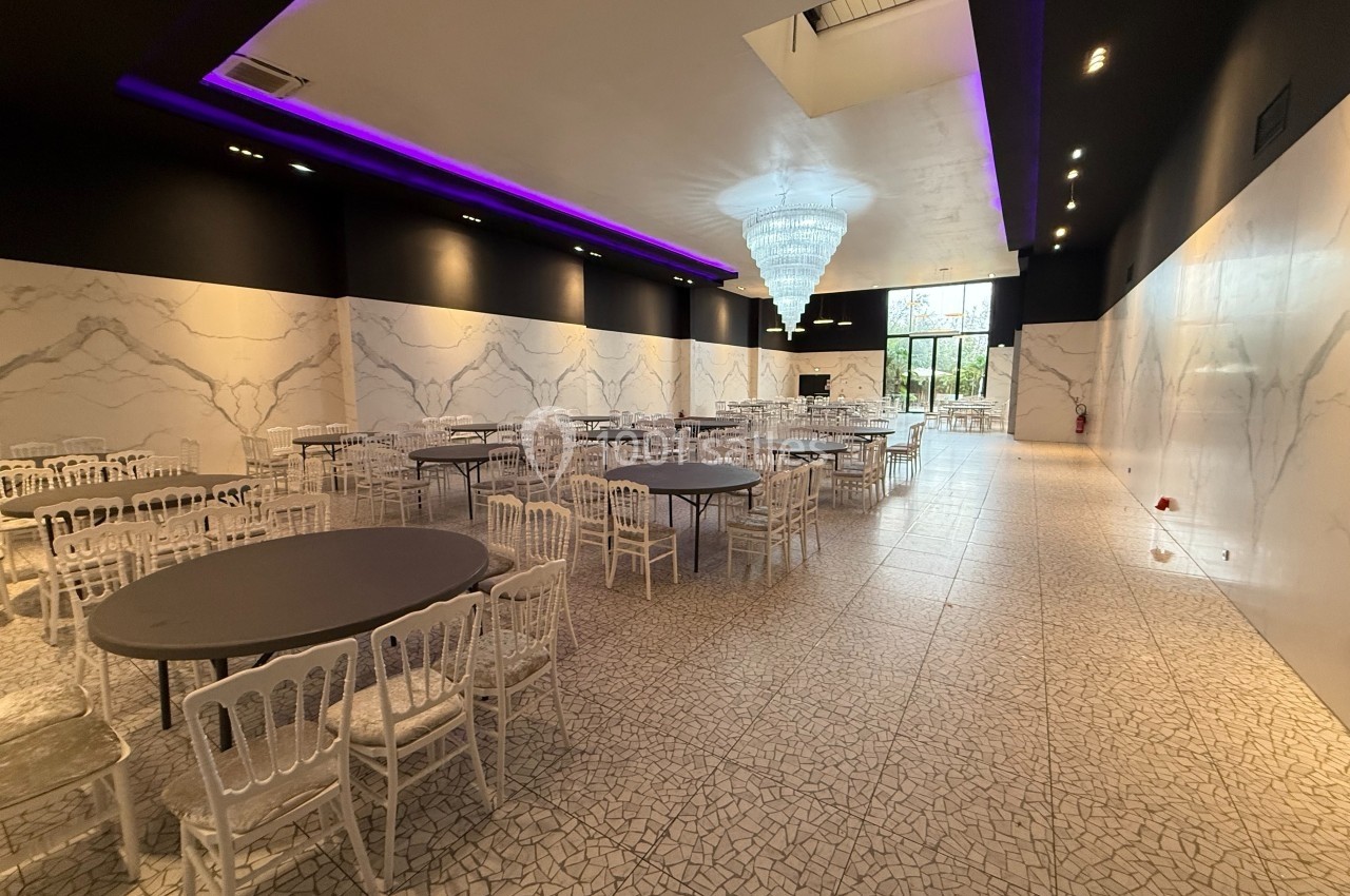 Salle villa Salle de réception spacieuse avec tables rondes, chaises blanches, murs effet marbre, lustre central et éclairage violet.