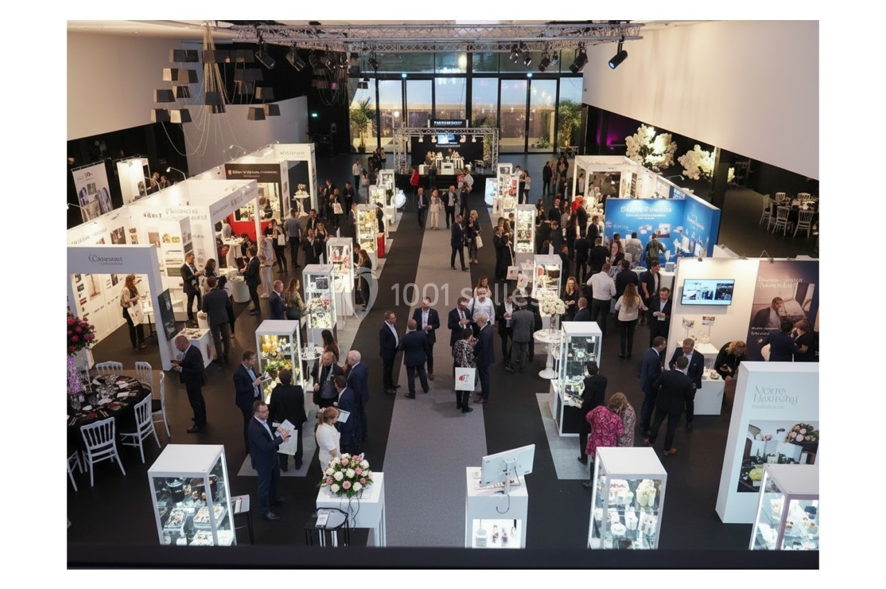 salon évènementiel Vue d'ensemble d'un salon professionnel avec des stands éclairés, des visiteurs et une grande baie vitrée en arrière-plan.