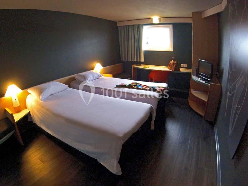 Chambre d'hôtel avec deux lits simples, bureau, télévision, éclairage tamisé et fenêtre avec rideaux.