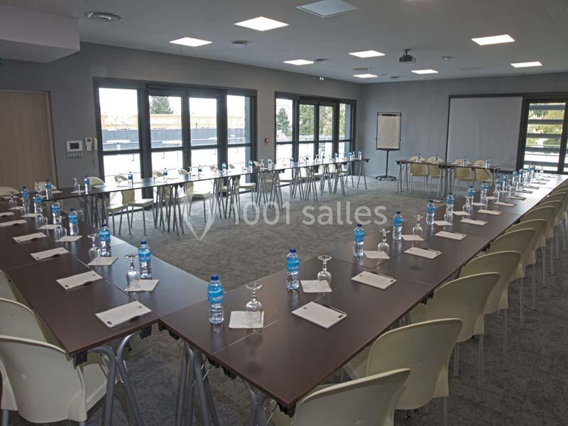 Salle de réunion lumineuse avec tables en U, chaises, bouteilles d'eau, verres et blocs-notes disposés pour les participants.