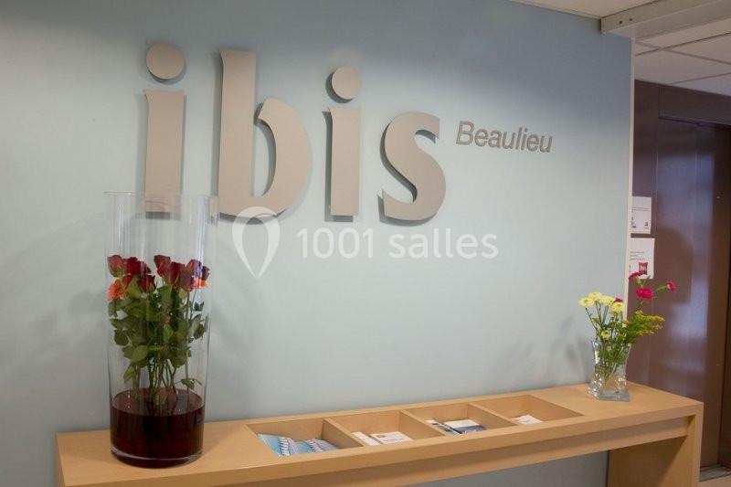 Logo ’ibis Beaulieu’ sur un mur bleu clair, avec un comptoir en bois décoré de vases contenant des fleurs.