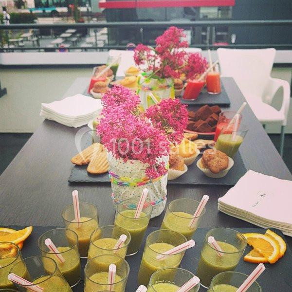 Table dressée avec des verrines de jus, muffins, biscuits et fleurs décoratives en extérieur.