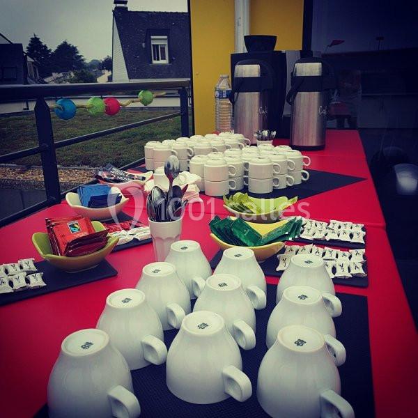 Table avec tasses, cuillères, sachets de thé, café et thermos, disposée pour un service de boissons en extérieur.