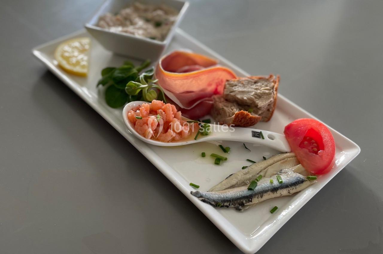 Assiette de dégustation avec crevettes, anchois, terrine, jambon, salade et tranche de tomate sur un plateau blanc.