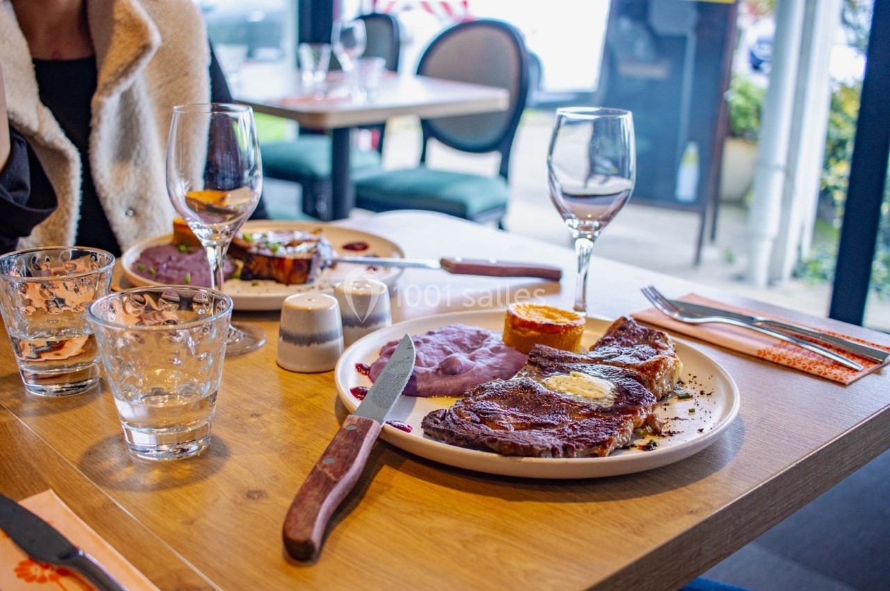 Assiette de viande grillée avec purée violette et accompagnements sur une table de restaurant, verres et couverts disposés.