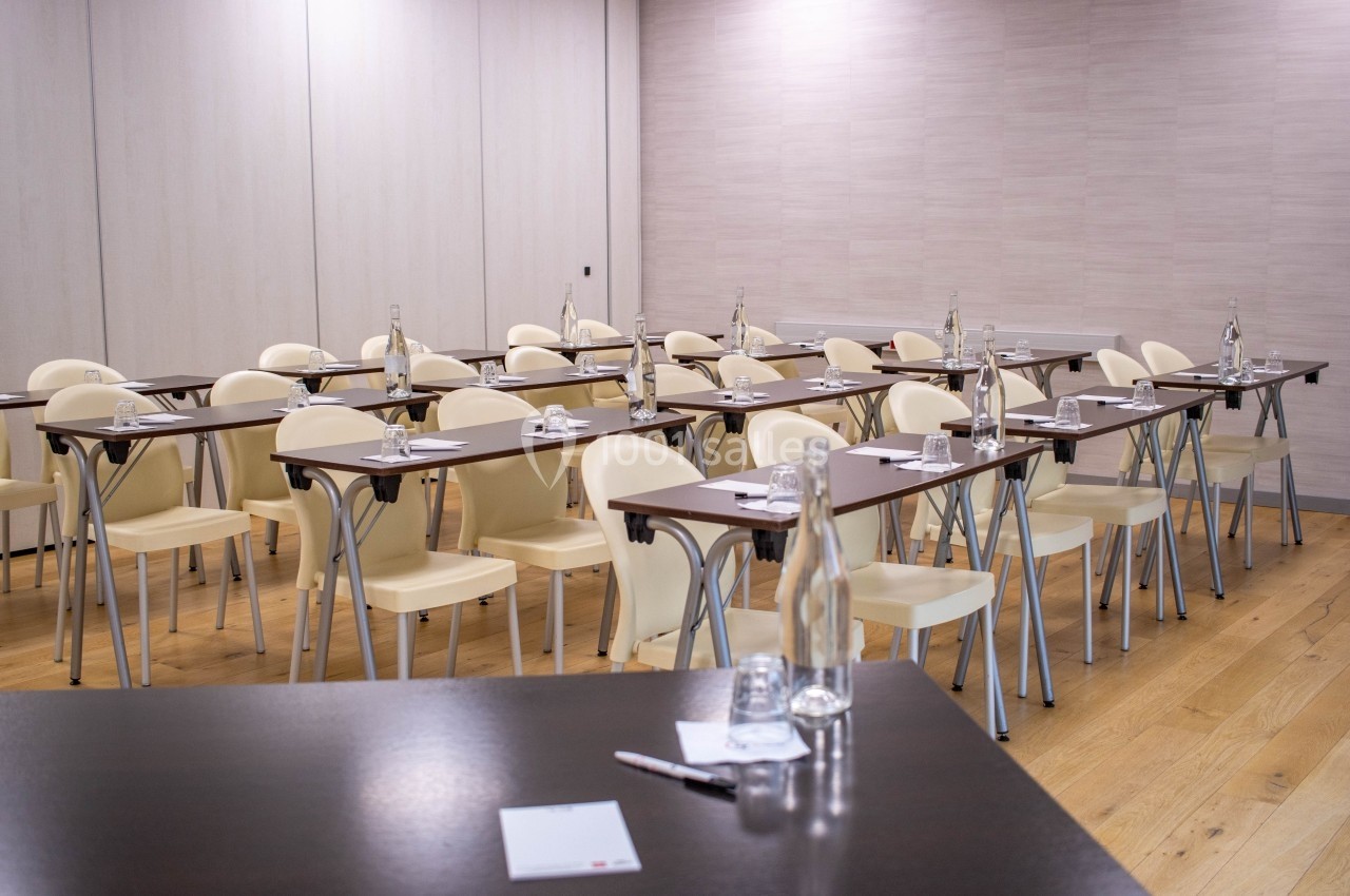Salle de réunion avec des tables alignées, chaises beiges, bouteilles d'eau et blocs-notes sur un sol en bois.