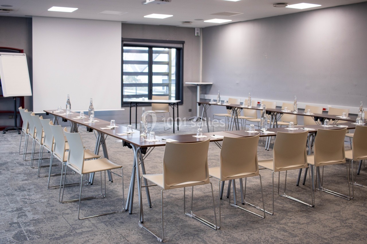 Salle de réunion lumineuse avec des tables disposées en U, des chaises, un tableau blanc et un écran de projection.