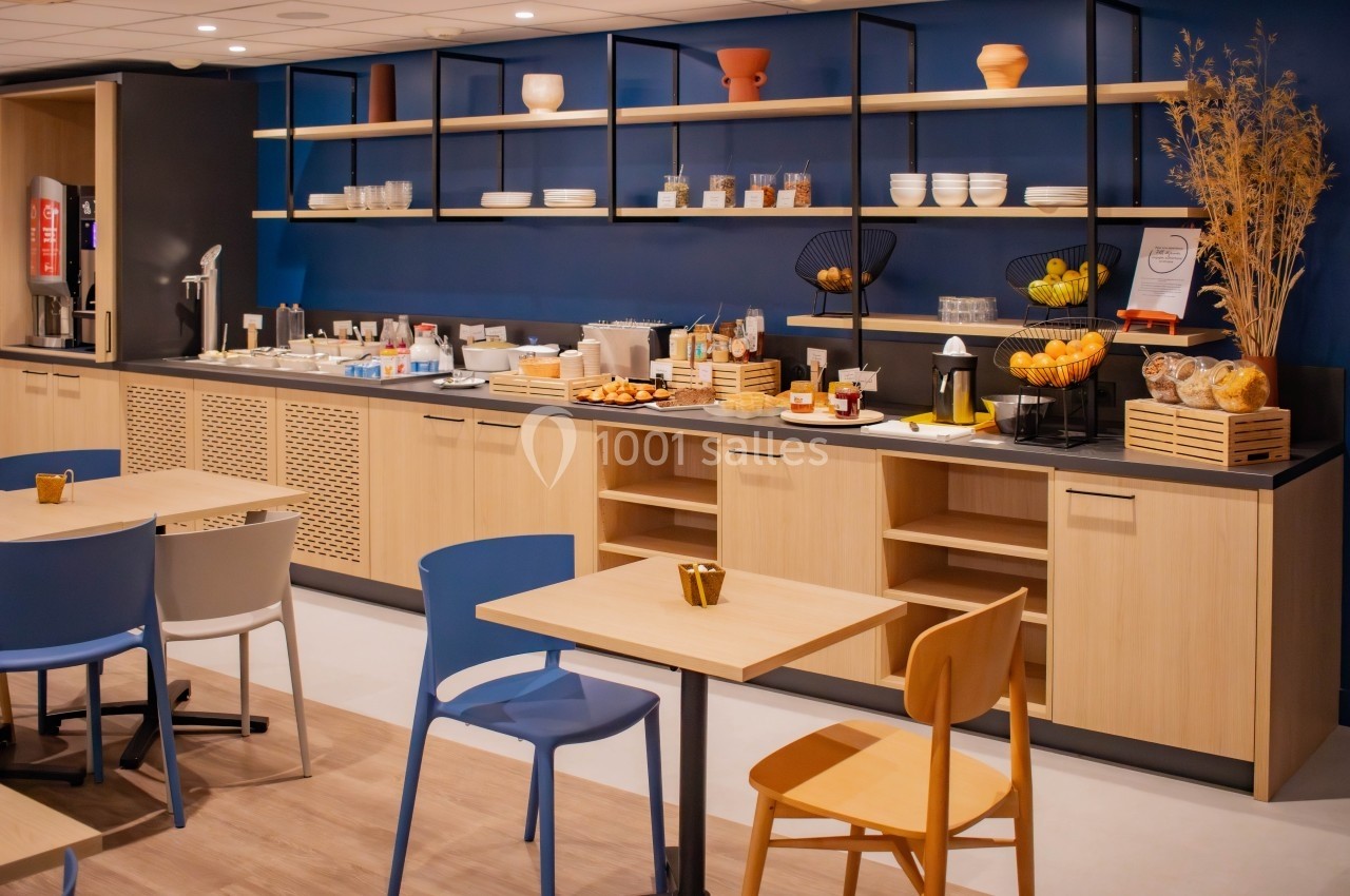 Buffet de petit-déjeuner avec boissons, fruits, viennoiseries et vaisselle, dans une salle aux tons bois et bleu.