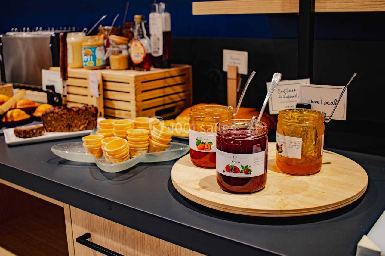 Pots de confiture et miel sur un plateau en bois, accompagnés de petits gaufrettes et pâtisseries en arrière-plan.