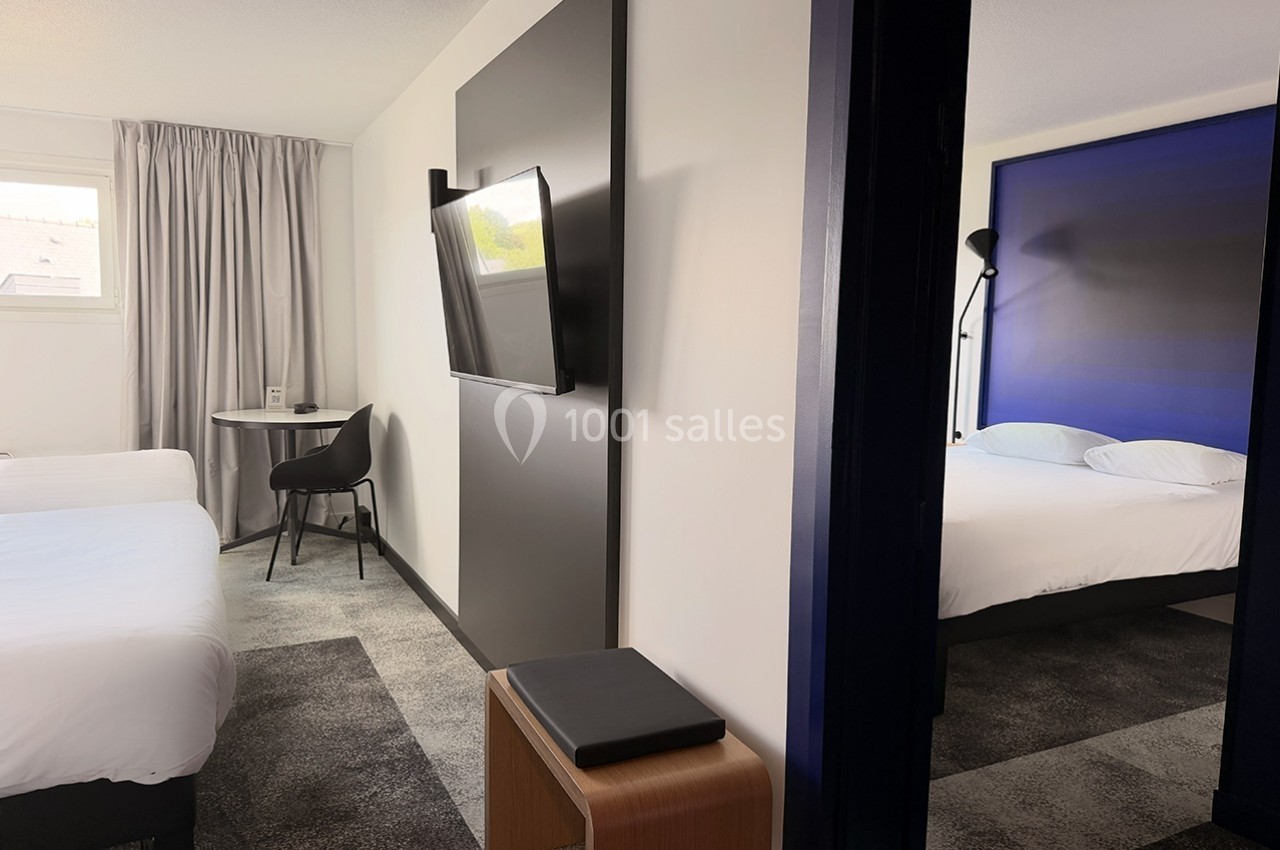 Chambre d'hôtel moderne avec deux lits simples, un bureau, une télévision murale et un aperçu d'une seconde pièce.